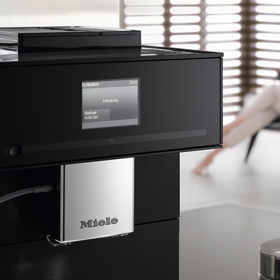 Schwarzer Miele Kaffeevollautomat mit silberner Blende und Display. Im Hintergrund Jalousien und eine Person.