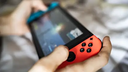 Eine Person hält eine schwarze Spielkonsole mit blauem und rotem Controller über einer hellen Decke.