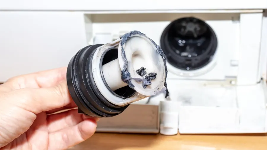 Hand hält einen verstopften, runden, weißen Waschmaschinenfilter mit schwarzen Rückständen vor einer weißen Waschmaschine.