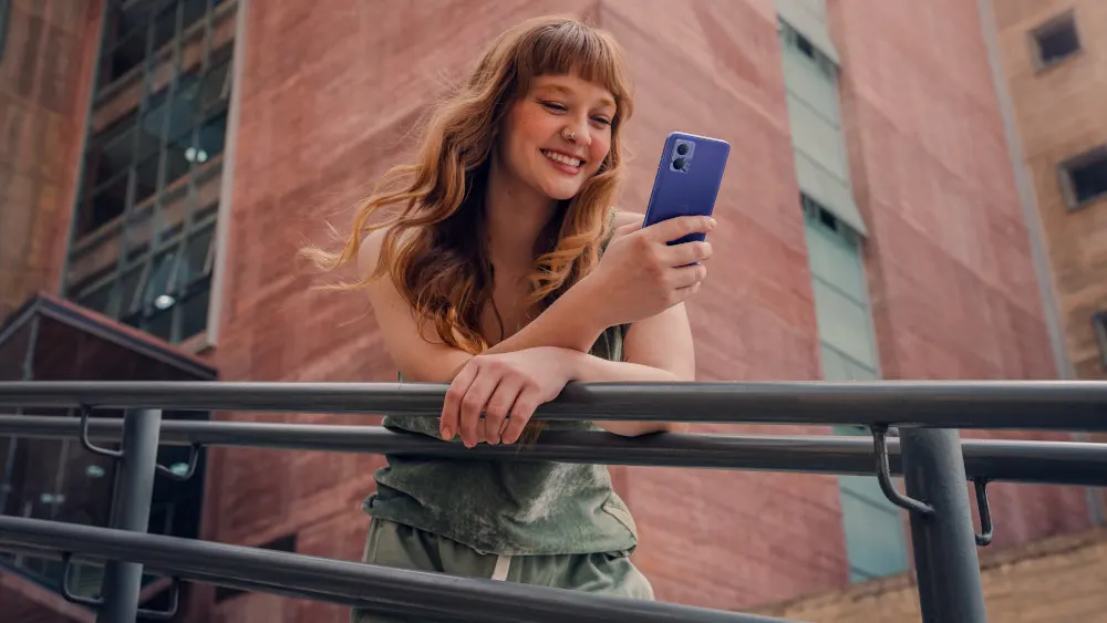 Eine Frau mit einem blauen Handy lehnt an einem grauen Geländer vor einem Backsteingebäude.