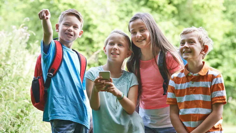 Vier Kinder im Freien. Einer zeigt, einer hält ein Smartphone, zwei lächeln. Bäume sind im Hintergrund.