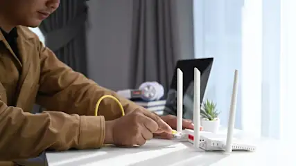 Ein Mann verbindet ein gelbes Ethernet-Kabel mit einem weißen Router mit drei Antennen auf einem weißen Tisch. Im Hintergrund sind ein Laptop, Kopfhörer und eine Pflanze zu sehen.