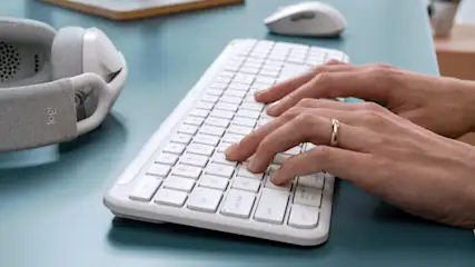 Hände tippen auf eine weiße Tastatur. Graue Kopfhörer, weiße Maus und ein Schreibtisch sind ebenfalls sichtbar.