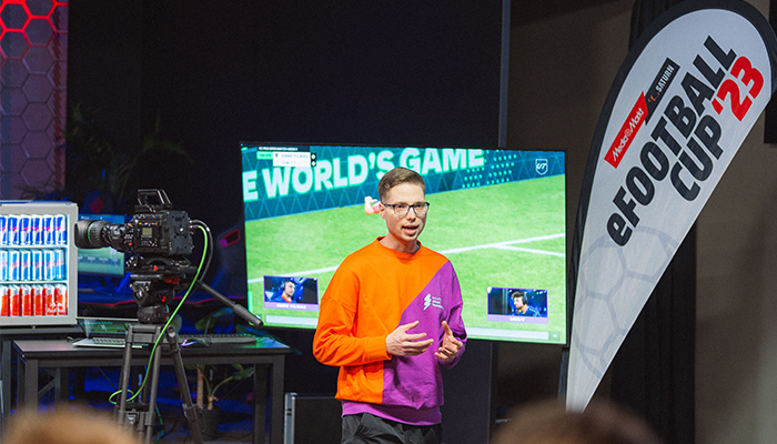 Ein Mann in einem orange-lila Pullover gestikuliert vor einem Bildschirm mit einem Fußballspiel. Eine Kamera und ein eFootball Cup '23 Banner sind sichtbar.