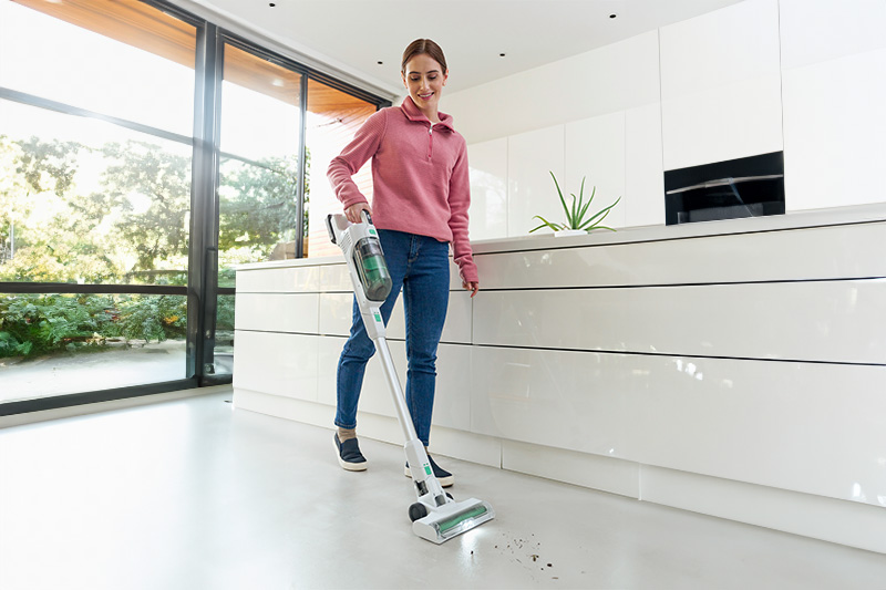 Eine Frau staubsaugt in einer Küche mit weißen Schränken und großem Fenster. Sie trägt Jeans und einen pinkfarbenen Pullover.