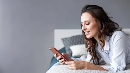 Eine Frau liegt auf einem Bett und hält ein rotes Mobiltelefon in ihren Händen. Im Hintergrund sind Kissen und eine graue Wand.