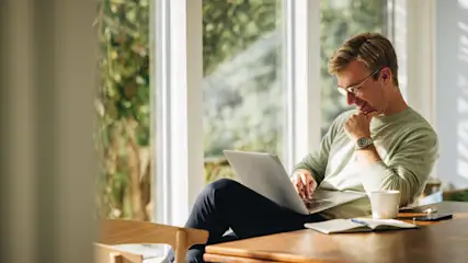 Ein Mann sitzt an einem Holztisch und benutzt einen Laptop, mit Blick auf ein Fenster und Grün im Hintergrund.