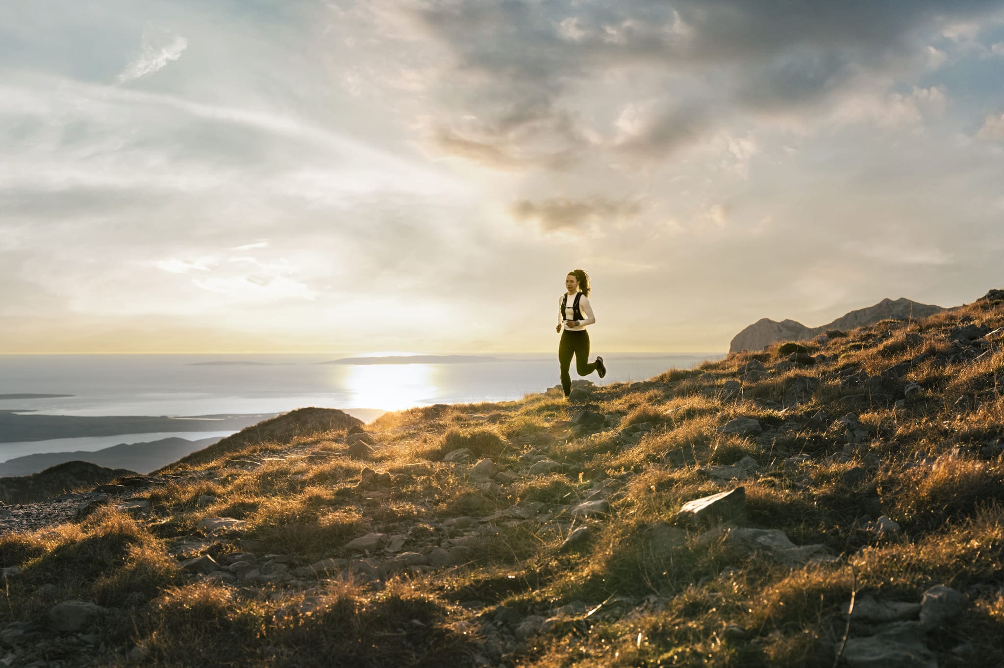  Trailrunnerin joggt bei Sonnenuntergang über einen felsigen Hang mit Meer und Bergen im Hintergrund.