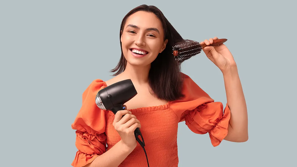 Eine Frau lächelt und hält eine schwarze Haarbürste im Haar und einen Fön in der Hand vor einem grauen Hintergrund.