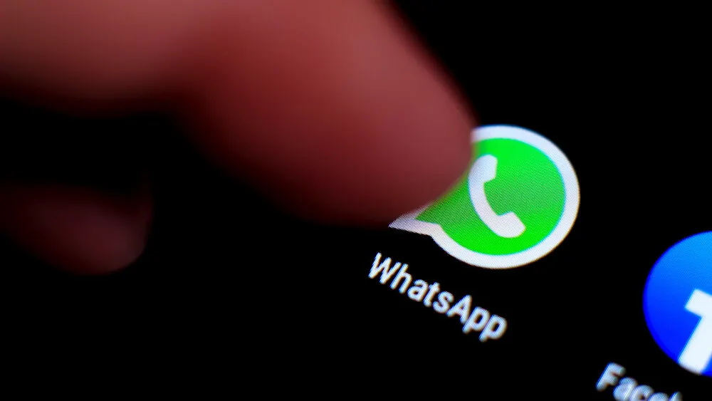 Nahaufnahme eines Fingers, der das grüne WhatsApp-Symbol auf einem schwarzen Smartphone-Bildschirm berührt. Ein blaues Facebook-Symbol ist teilweise sichtbar.