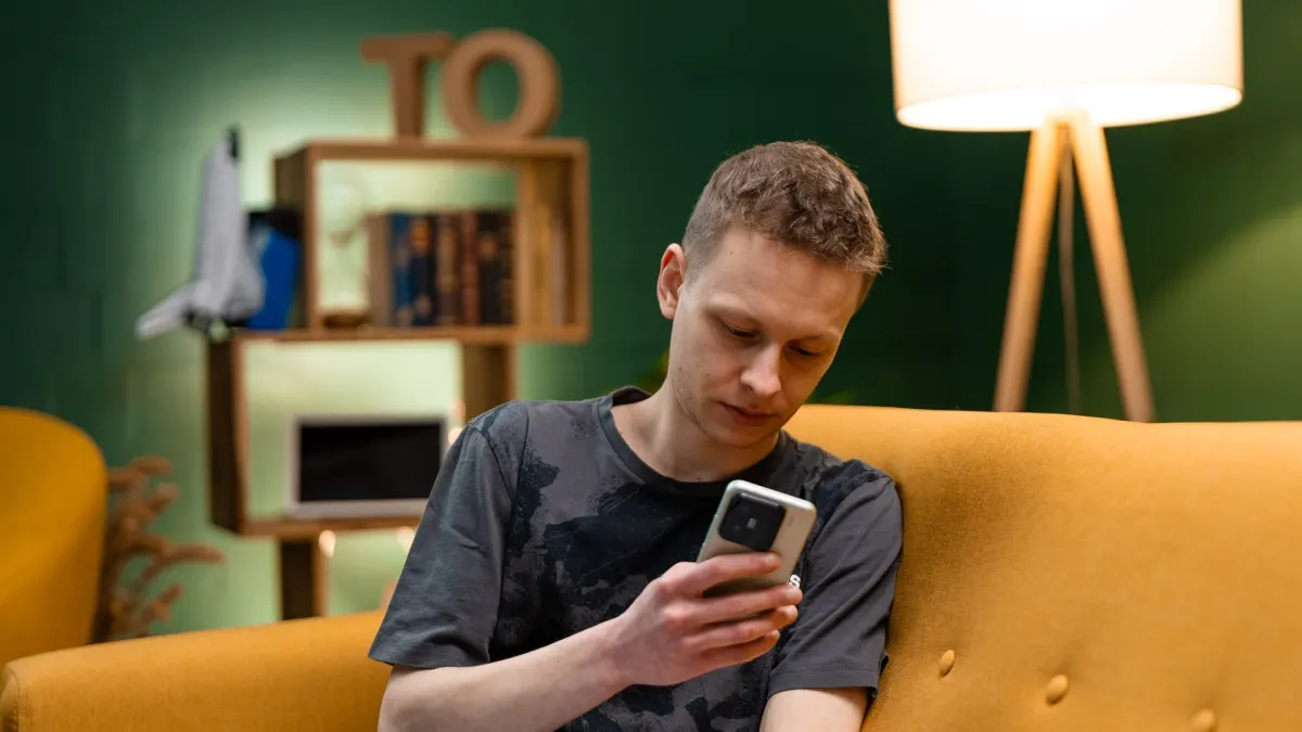 Ein Mann sitzt auf einem gelben Sofa und schaut auf sein Handy; im Hintergrund befinden sich ein Regal und eine Stehlampe.