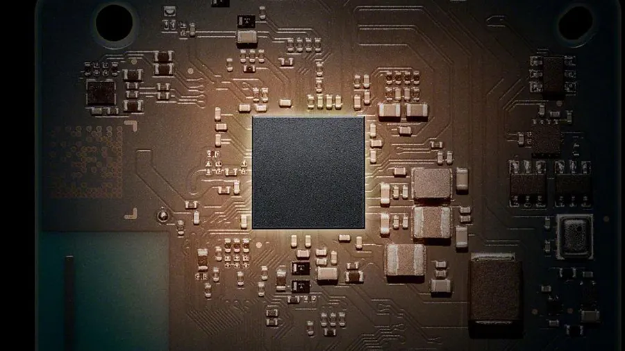 Eine Nahaufnahme einer braunen Leiterplatte mit einem quadratischen, schwarzen Chip in der Mitte, umgeben von elektronischen Bauteilen.