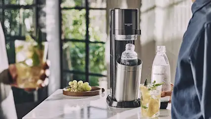 Ein Sodastream-Gerät steht auf einem Marmortisch, daneben eine Flasche, ein Glas mit Getränk und eine Schale mit Weintrauben.