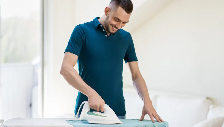 Ein Mann in einem blauen Poloshirt bügelt ein hellblaues Tuch auf einem Bügelbrett mit einem weißen Bügeleisen.