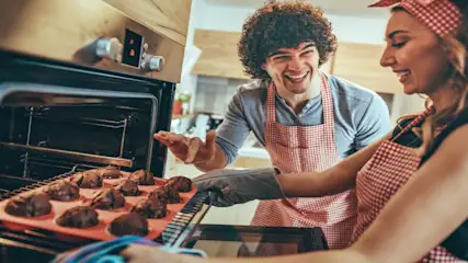 Ein Mann und eine Frau mit rot-weiß karierten Schürzen lachen neben einem Ofen mit Muffins.