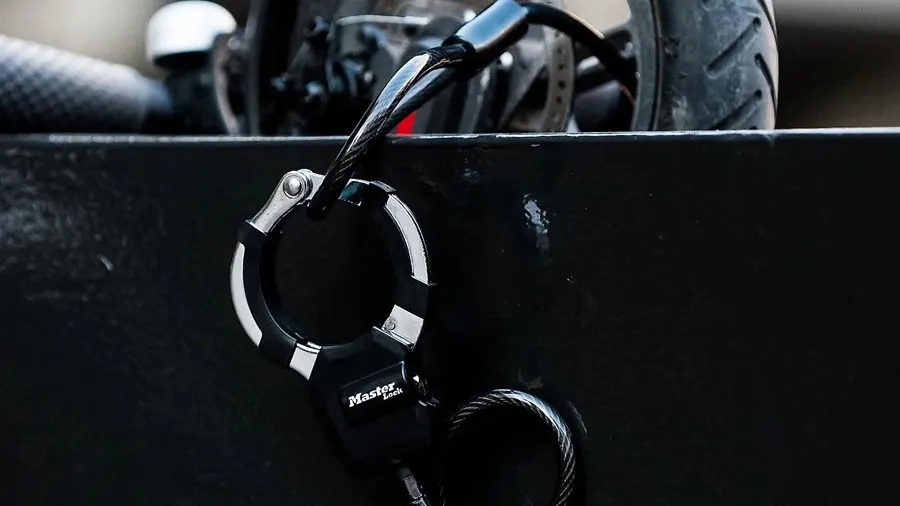 MASTERLOCK 66268700 Street Cuff Ein schwarzes Master Lock-Fahrradschloss sichert ein Motorrad mit schwarzem Kabel an einem schwarzen Hintergrund.