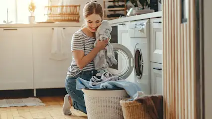 Eine Frau kniet vor einer Waschmaschine, hält ein Stofftier und hat einen Wäschekorb vor sich.
