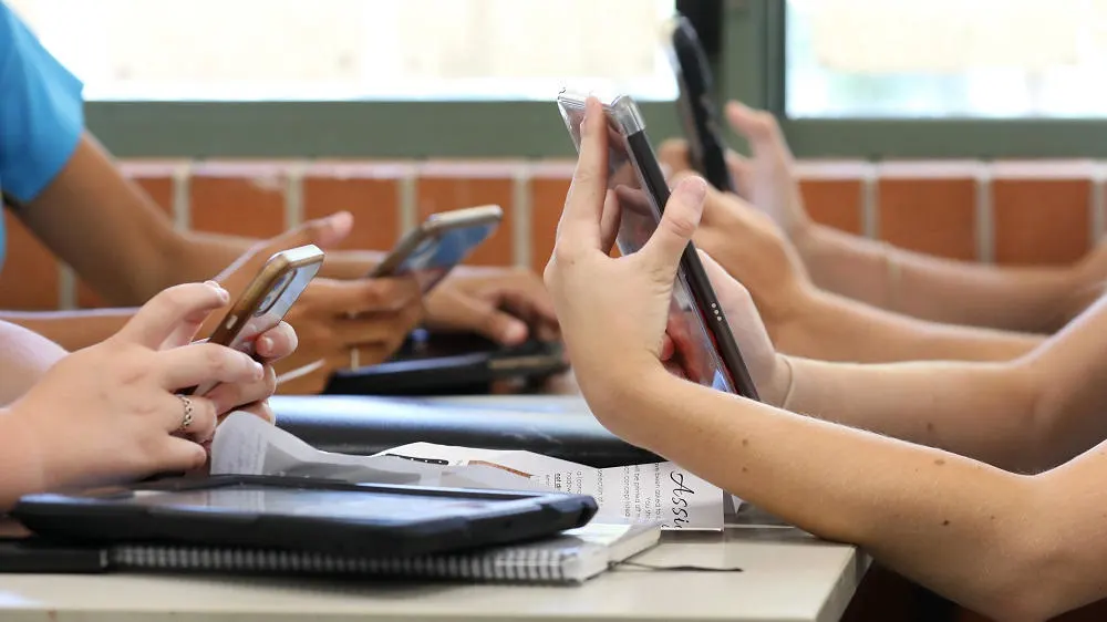 Personen sitzen am Tisch und halten Handys. Tablets, Notizbücher und Papiere liegen auf dem Tisch.