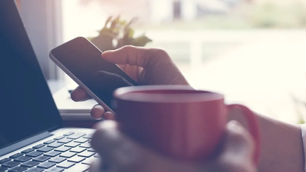 Eine Person tippt auf einem Smartphone, während sie eine rote Tasse hält, mit einem Laptop und Fenstern im Hintergrund.