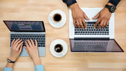 Zwei Personen tippen auf Laptops. Zwei Tassen Kaffee stehen auf einem hellen Holztisch.