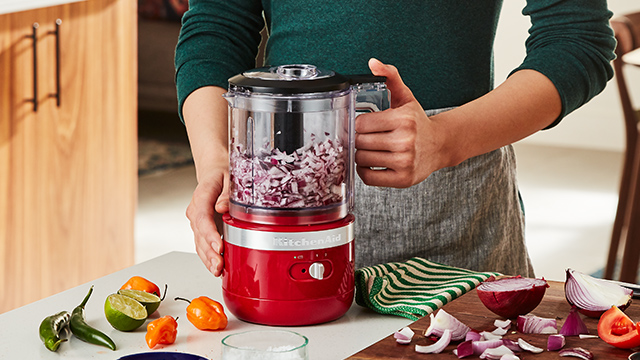 Eine Person hält einen roten Küchenmixer mit gehackten Zwiebeln. Auf dem Tisch liegen Limetten, Chilis und eine Holzplatte mit Zwiebeln.