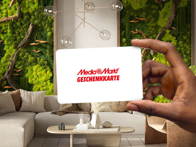 Eine Hand hält eine weiße MediaMarkt Geschenkkarte mit rotem Schriftzug ins Bild vor dem Hintergrund eines Wohnzimmers mit Pflanzen.