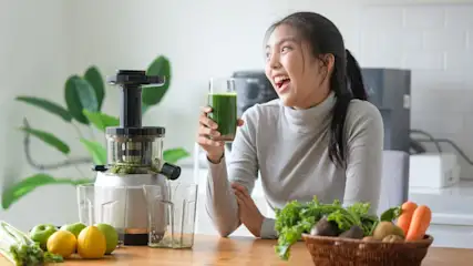 Eine lachende junge Frau steht mit einem Glas grünen Saft neben einem Slow Juicer in einer Küche. (Adobe Stock Bildnr.: 1569541961)