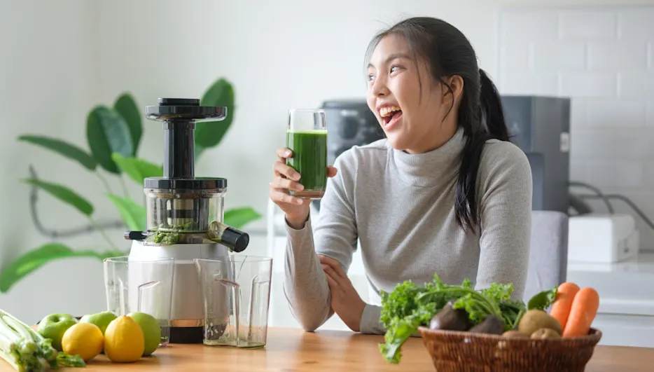 Eine lachende junge Frau steht mit einem Glas grünen Saft neben einem Slow Juicer in einer Küche. (Adobe Stock Bildnr.: 1569541961)