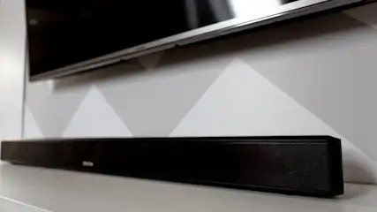 Ein schwarzer Fernseher hängt über einer schwarzen Soundbar auf einem weißen Tisch vor einer Wand mit grauen Dreiecken.