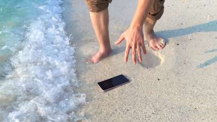 Handy liegt am Strand auf dem Boden. Eine kleine Welle nähert sich. Eine Männerhand greift danach.