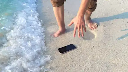 Eine Person steht barfuß am Strand neben einem schwarzen Handy, während Wellen an den Strand rollen.