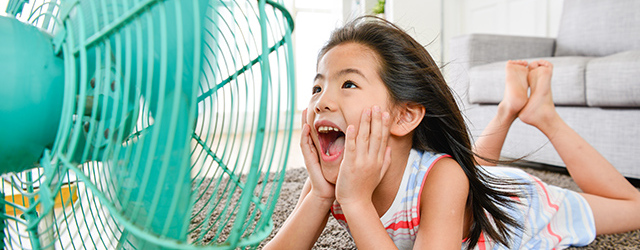 Ein Mädchen liegt auf einem Teppich und freut sich über einen grünen Ventilator, der neben ihr steht. Im Hintergrund steht ein graues Sofa.