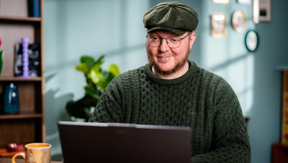 Mann mit Brille und Schirmmütze sitzt vor einem Laptop. Im Hintergrund sind ein Regal und eine Pflanze zu sehen.