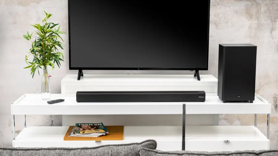 Ein Fernseher steht auf einem weißen Regal mit einer Soundbar und einem schwarzen Lautsprecher. Eine Pflanze steht links daneben.