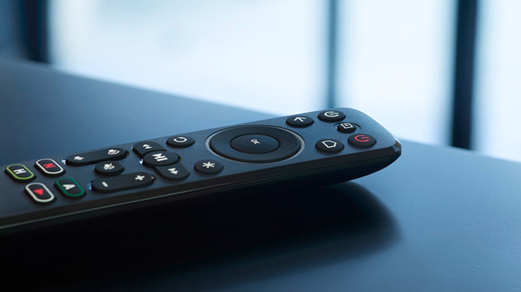Eine schwarze Fernbedienung mit Knöpfen liegt auf einer blauen Oberfläche vor einem verschwommenen, hellen Hintergrund.