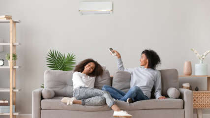 Zwei Frauen sitzen im Wohnzimmer auf dem Sofa und eine von ihnen schaltet eine Split-Klimaanlage ein.