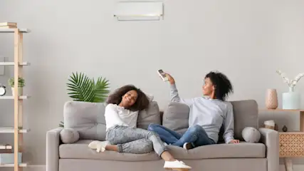 Zwei Frauen sitzen auf einem grauen Sofa unter einer Klimaanlage und eine hält eine Fernbedienung.