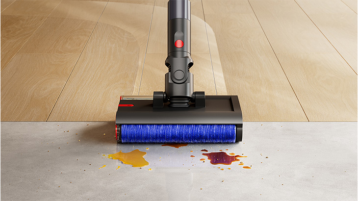 Ein moderner kabelloser Staubsauger mit blauer Bürstenrolle reinigt eine Übergangsfläche zwischen hellem Holzboden und grauem Betonboden. Auf dem Beton sind gelbe und rote Flüssigkeitsflecken sichtbar, die vom Staubsauger gereinigt werden.