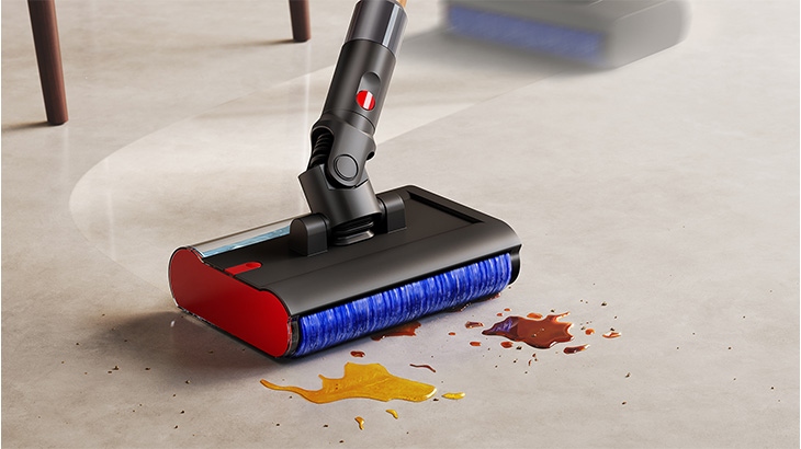 Nahaufnahme eines modernen kabellosen Staubsaugers mit roter und schwarzer Bürstenwalze auf hellem Betonboden. Die Bürste mit blauen Borsten reinigt gelbe und rote Flüssigkeitsflecken. Im unscharfen Hintergrund sind Möbelbeine sichtbar.
