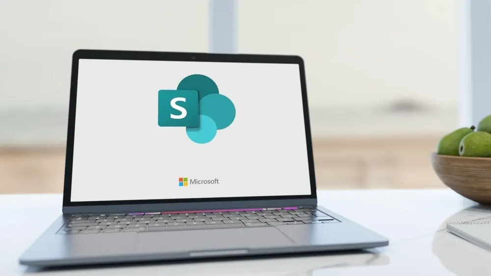Ein Laptop zeigt das Microsoft SharePoint-Logo auf dem Bildschirm. Auf einem Tisch stehen eine Schale mit grünen Früchten und ein Notizbuch.