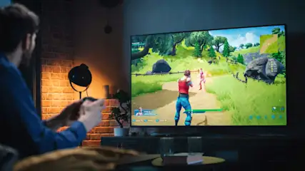 Mann spielt Videospiel auf großem Bildschirm in einem Wohnzimmer. Eine Lampe steht neben einer Ziegelwand. Zimmerpflanze auf dem Tisch.