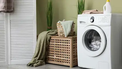 Ein weißer Wäschetrockner steht neben einem Holzschrank mit Wäschekorb in einem Waschraum.