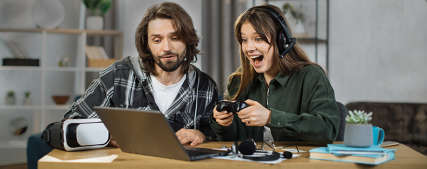 Eine aufgeregte, fokussierte junge Frau mit Headset, die an einem Laptop ein Computerspiel spielt. Daneben sitzt ein junger Mann, der von ihrer Leistung beeindruckt ist.