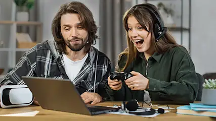 Ein Mann und eine Frau sitzen an einem Tisch mit Laptop, VR-Brille, Controller und Kopfhörern. Die Frau trägt ein Headset und freut sich.