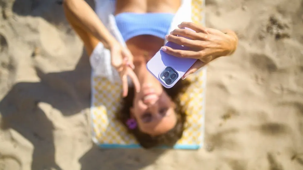Eine Person liegt auf einem gelb-weiß karierten Tuch am Strand und hält ein lila Handy.