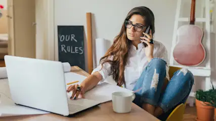 Eine Frau mit Brille telefoniert am Schreibtisch mit Laptop, Notizen und Tasse. Im Hintergrund Gitarre und Tafel.
