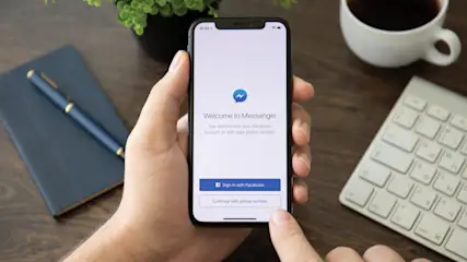 Ein Mobiltelefon zeigt den Messenger-Anmeldebildschirm, gehalten von einer Hand. Ein Notizbuch, ein Stift, eine Kaffeetasse, eine Pflanze und eine Tastatur liegen auf dem Tisch.