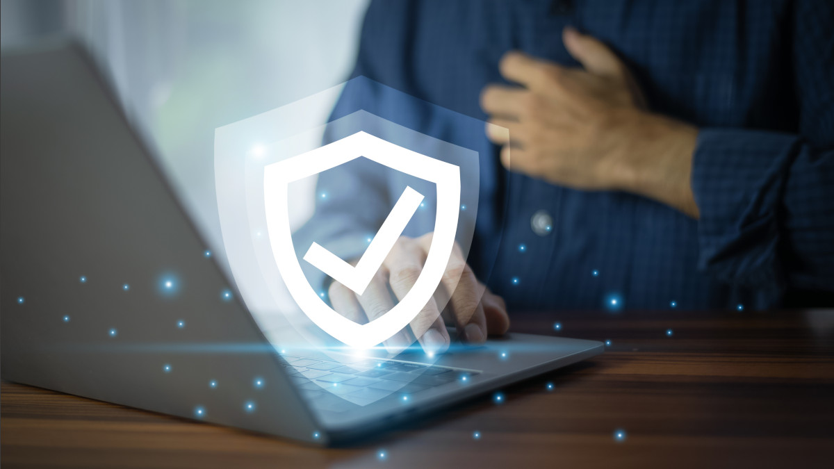 Eine Person tippt auf einem Laptop. Ein weißes Schildsymbol mit Häkchen liegt darüber, blaue Punkte umgeben den Laptop.
