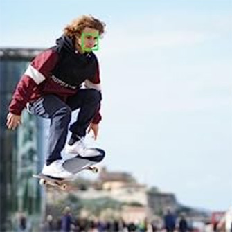 Ein Skateboarder in der Luft, bekleidet mit einer rot-schwarzen Jacke und blauen Hosen, vor einem verschwommenen städtischen Hintergrund.