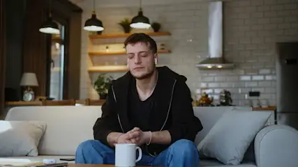 Ein Mann sitzt mit geschlossenen Augen auf einem Sofa mit Kopfhörern. Vor ihm steht eine Tasse. Im Hintergrund sind Küchengeräte.